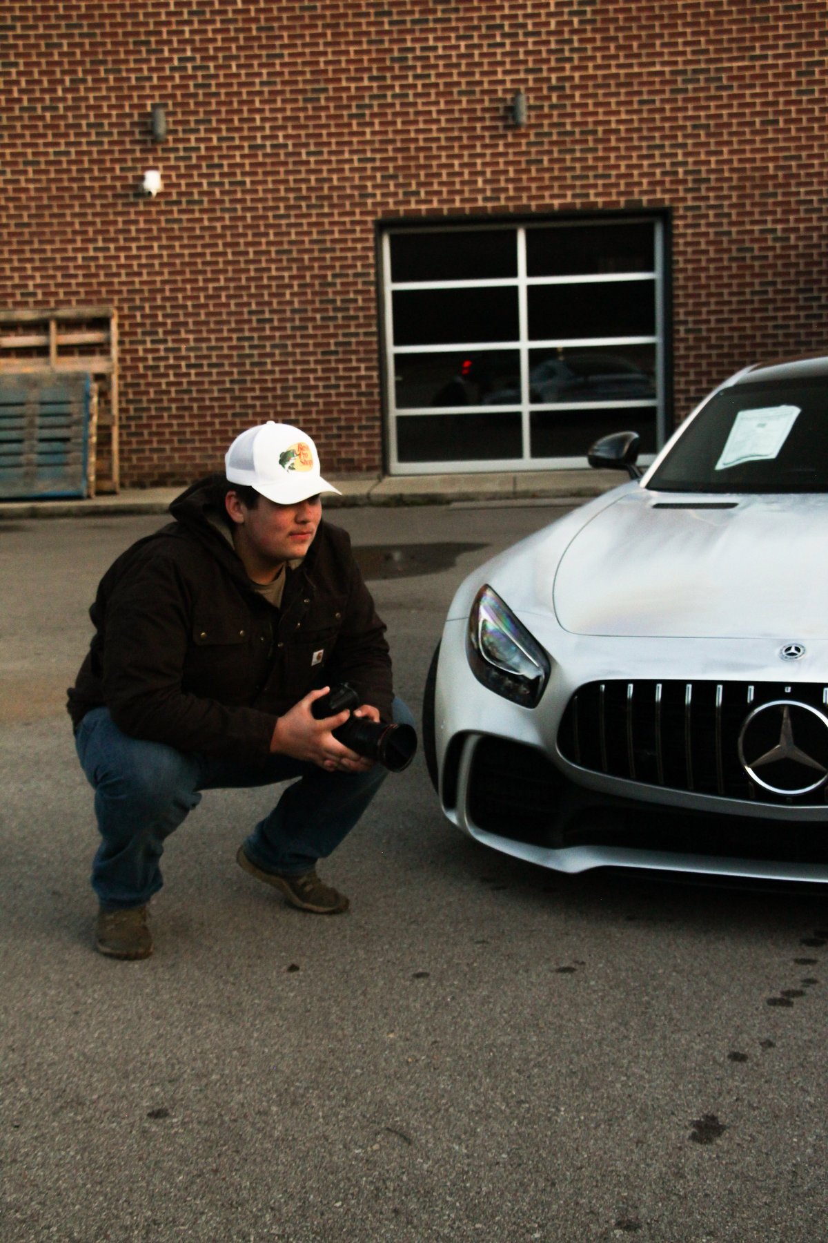 Man in white cap and black jacket crouching beside a silver Mercedes sports car in a parking lot with brick building behind